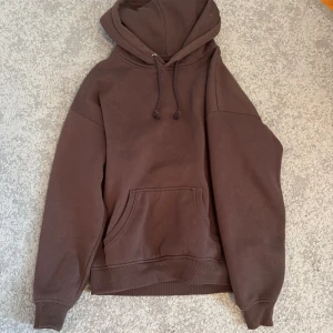 Brun hoodie - Säljer en brun hoodie med en klassisk design. Den har en stor ficka framtill och justerbar huva med dragsko. Perfekt för en avslappnad stil.