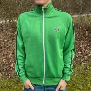 Grön track jacket från Fred Perry - Säljer en grön track jacket från Fred Perry med vita och svarta ränder längs ärmarna. Jackan har en dragkedja framtill och ribbade muddar vid ärmslut och nederkant. Perfekt för en sportig look!