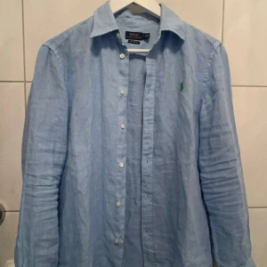 Blå linneskjorta från Ralph Lauren - Snygg blå linneskjorta från Ralph Lauren med relaxed fit. Skjortan har långa ärmar och knappar framtill. Den är broderad med det klassiska gröna Polo-loggan på bröstet. Perfekt för en stilren look! Sjukt bra pris, endast 200kr