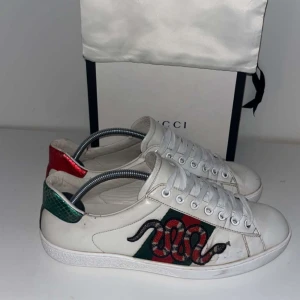 Vita sneakers med ormmönster - Snygga vita sneakers med ett coolt ormmönster på sidan. Skorna har snörning och en stilren design med röda och gröna detaljer på hälen. Perfekta för en trendig look.