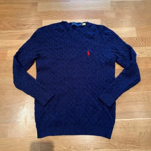 Blå stickad tröja från Ralph Lauren - Säljer en snygg blå stickad tröja från Ralph Lauren med det klassiska röda logotypen på bröstet. Tröjan har ett kabelstickat mönster och långa ärmar. Perfekt för kyligare dagar! Storlek M, endast använd ett fåtal gånger.