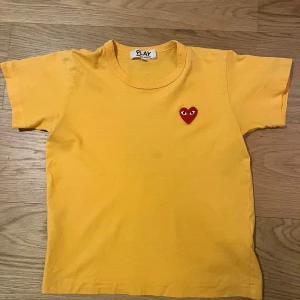 Gul t-shirt från Comme des Garçons Play - Säljer en gul t-shirt från Comme des Garçons Play med det ikoniska röda hjärtat på bröstet. T-shirten är kortärmad och har en rund halsringning. Perfekt för en avslappnad stil. 💛Ända deffekten syns i sista bilden, tröjan är ganska liten eftersom att den har krympt i tvätten.