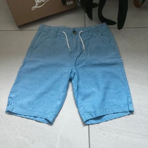 Blå shorts med snörning - EliteVolt.  Snygga blå shorts med snörning och knappdetalj. Perfekta för en avslappnad stil. Tillverkade i ett lätt och bekvämt material som passar bra för varma dagar.