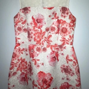 Blommig klänning med spetsdetaljer - Charmig klänning med blommigt mönster i rött och rosa på en vit bakgrund. Den har en vacker spetsdetalj upptill och en figurnära passform. Perfekt för somriga tillfällen.