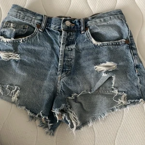 Blå jeansshorts från Zara - Säljer ett par coola blå jeansshorts från Zara med slitna detaljer och fransiga kanter. Shortsen har en klassisk femficksdesign och knappgylf. Perfekta för en avslappnad stil!