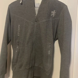 Grå hoodie med dragkedja från IFK Göteborg - Säljer en mkt sällsynt grå hoodie med dragkedja från IFK Göteborg. Tröjan har en broderad lejon logga på bröstet och texten 'Göteborg' längs sidan. Den har en Luva och två fickor med dragkedja framtill. Köpt för 1149 och är i bra skick utan defekter. Tröjan passar även S. Kan tänka mig gå ner i pris vid snabb och smidig affär! Skriv om du har några funderingar.