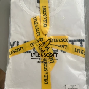 Vit t-shirt och blå-rutig byxa från Lyle & Scott - Säljer ett set med en vit t-shirt och blå-rutiga byxor från Lyle & Scott. T-shirten har en klassisk crew neck och byxorna är i ett bekvämt lounge-stil. Perfekt för en avslappnad dag hemma. Setet är snyggt förpackat med ett gult band med Lyle & Scott-loggan. Sköna mjuka matchande byxor tillkommer. Nypris i butik: 689 ellos. 