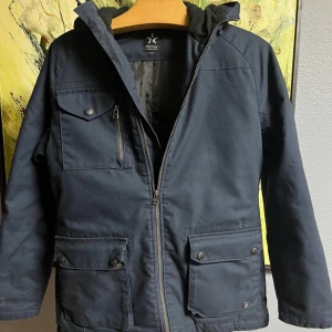 Texstar Jacka - Säljer en mörkblå jacka från Texstar med huva och flera praktiska fickor. Jackan har dragkedja och knappar framtill samt en stilren design. Perfekt för våren! Jackan är i väldigt bra skick och inga tecken på användning. 