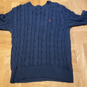 Ralph Lauren knit  - Säljer en stilren mörkblå kabelstickad tröja från Polo Ralph Lauren. Tröjan har en klassisk design med ett rött broderat märke på bröstet och ribbade kanter vid ärmslut. Skriv gärna vid minsta lilla funderingar 