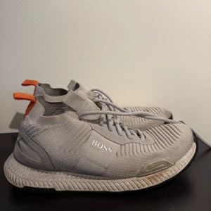 Grå sneakers från Boss - Snygga grå sneakers från Boss med stickad design och snörning. Skorna har en modern look med orange detaljer vid hälen och en bekväm sula.