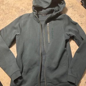 Grå hoodie från Sail Racing - Säljer en stilren grå hoodie från Sail Racing med dragkedja och broderad logga på bröstet. Tröjan har en praktisk huva och långa ärmar, perfekt för kyliga dagar. Den är tillverkad i ett mjukt material som ger en skön känsla. Passar både till vardags och mer avslappnade tillfällen.