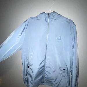 Ljusblå vindjacka från Trapstar - Säljer en stilren ljusblå vindjacka från Trapstar med dragkedja och huva. Jackan har en cool logga på bröstet och text på ryggen. Perfekt för att hålla vinden borta med stil. 🌀