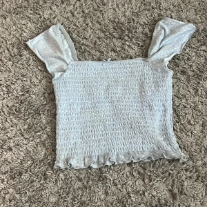 Ljusblå smockad topp med volang - Supersöt ljusblå topp med smockad kropp och volangkant nertill. Axelbanden är breda och har ett diskret vitt prickigt mönster som ger en gullig touch. Perfekt för dig som gillar en feminin och trendig stil.