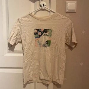 Beige t-shirt med kattmotiv från Uniqlo - Beige t-shirt från Uniqlo UT i bomull med ett färgglatt tryck av en söt katt och blommor på bröstet. Klassisk passform med rund hals och korta ärmar. Perfekt för dig som gillar unika prints och vill sticka ut lite extra.