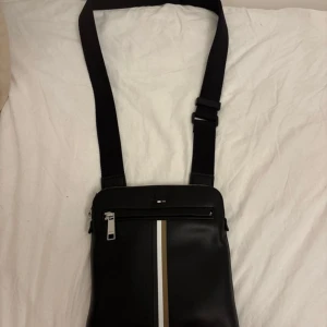 Boss axelväska i läder - Snygg svart axelväska från BOSS i skinn med justerbar rem. Man får gympakassan med, de finns 3 platser att lägga saker i väskan. Perfekt för att lägga in sin lur och laddare o massor mer där inne, hör av er så fort ni kan! 