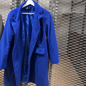 Kornblå kappa med klassisk krage - Snygg kornblå kappa med klassisk krage och fickor framtill. Jackan har en rak passform och är helfodrad för extra komfort. Perfekt för dig som vill sticka ut med färg i höst och vinter. Stängs med knappar framtill.