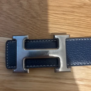 Mörkblått dubbelsidigt skärp med H-spänne - Säljer ett stilrent marinblått skärp med ikoniskt H-spänne i silverfärgad metall. Bältet är gjort i präglat skinn med vita kontrastsömmar och klassisk design. Perfekt för dig som vill ha en lyxig accessoar med statement-känsla.