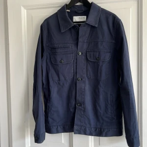 Marinblå overshirt från Selected Homme - Snygg marinblå overshirt från Selected Homme med klassisk krage, knäppning framtill och två bröstfickor med lock och knapp. Tillverkad i bomull, perfekt för lager-på-lager och stilren look. Storlek L men liten i storleken.