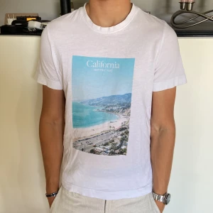 Vit t-shirt med California-tryck - Vit t-shirt med ett stort färgglatt tryck framtill som visar en strand och texten 'California summer surf'. Klassisk rund halsringning och korta ärmar. T-shirten har en avslappnad passform och är gjord i mjukt bomullstyg. Perfekt för dig som gillar chill och somriga vibes. Funkar även för dig som har storlek S