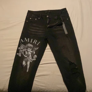 Svarta Amiri jeans med ängeltryck - Svarta jeans från Amiri med stort vitt ängeltryck och logga på vänster ben. Jeansen har slitna detaljer och hål på höger knä, klassisk femficksdesign och läderpatch bak i midjan. Snygg streetstil med smal passform och coola detaljer.