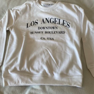 Vit sweatshirt med Los Angeles-tryck - Vit sweatshirt med svart texttryck på framsidan. Tröjan har rund hals, långa ärmar och ribbade muddar vid ärmslut och nederkant. Perfekt för dig som gillar streetstyle och enkel design.