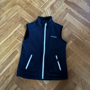 Mörkblå väst från Rosett Riding - Sportig mörkblå väst från Rosett Riding med vit dragkedja framtill och två fickor med vita dragkedjor. Hög krage och diskret logga på bröstet. Perfekt för ridning och utomhusaktiviteter, tillverkad i mjukt syntetmaterial.