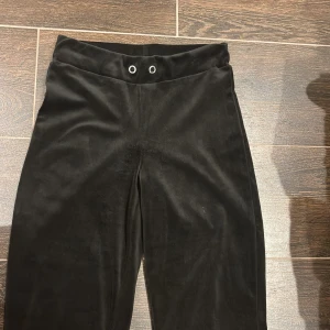 Svarta sammetsbyxor från Gina Tricot - Svarta byxor i mjuk sammet från Gina Tricot. Modellen har hög midja med två metallöljetter framtill och raka ben. Perfekta för dig som gillar en stilren look med lite extra lyxig känsla tack vare materialet.