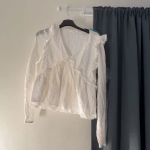 Vit volangblus med v-ringning - Supersöt vit blus med v-ringning och volangdetaljer. Långa ärmar och lätt, luftig känsla. Perfekt för dig som gillar romantisk och bohemisk stil. Sitter snyggt och har fina detaljer vid axlarna.