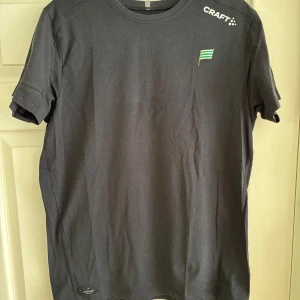 Svart hammarbytröja - Svart t-shirt från Craft med liten grön och vit flagga på bröstet och logga på axeln. Klassisk rund hals och korta ärmar. Perfekt för dig som gillar enkel och sportig stil.