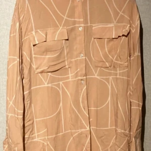 Beige mönstrad skjorta Visual Clothing Project - Snygg beige skjorta från Visual Clothing Project med abstrakt mönster i vitt. Skjortan har två stora bröstfickor med lock, knäppning framtill och långa ärmar. Perfekt för dig som gillar en avslappnad men trendig stil.