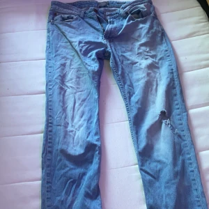Blå jeans med slitning på benet - Säljer ett par klassiska blå jeans med snygg slitning på ena benet. Jeansen har fem fickor, normal passform och raka ben. Perfekta för en avslappnad streetstil och tillverkade i mjukt jeanstyg.