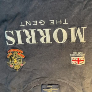 Mörkblå Morris Gents t-shirt M - Snygg mörkblå t-shirt från Morris Gents med broderade detaljer och tryck på bröstet, inklusive brittisk flagga och emblem. Stor text 'Morris The Gent' framtill och siffran 68 på ärmen. Klassisk passform och rund hals. Perfekt för dig som gillar preppy stil.