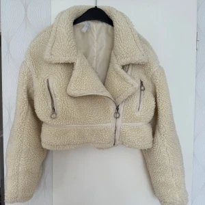 Beige croppad teddyjacka med dragkedjor - Säljer en beige croppad teddyjacka med coola silverfärgade dragkedjor och stora krage. Jackan har lång ärm och är gjord i mjukt, fluffigt teddy-material som håller dig varm och ger en trendig look. Perfekt för dig som vill sticka ut i höst och vinter.