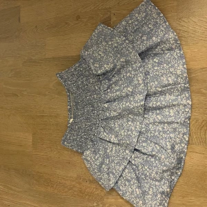 Blommig blå volangkjol Gina Tricot - Supersöt blå kortkjol från Gina Tricot med vitt blommigt mönster. Kjolen har bred smock i midjan och två lager volanger