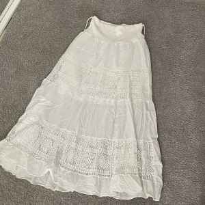 Vit långkjol med spetsdetaljer - Supersöt vit långkjol med bred linning och flera lager. Kjolen har inslag av spets och broderade blommor i mönstret, vilket ger en romantisk och bohemisk vibe. Perfekt för varma dagar och chill häng. Materialet känns lätt och luftigt.