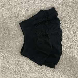 Svart volangkjol med resårmidja - Supersöt svart kortkjol med flera lager volanger och bred resår i midjan. Perfekt för dig som gillar en lekfull och trendig stil. Kjolen är enkel att matcha och har en loose passform som ger skön rörelsefrihet.