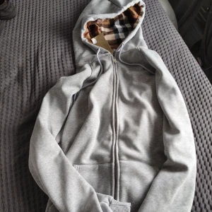 Grå Burberry hoodie med rutig huva - Säljer en grå hoodie från Burberry med dragkedja och snörning. Insidan av huvan har det klassiska Burberry-rutiga mönstret i beige, brunt och svart. Tröjan har långa ärmar och två fickor framtill. Perfekt för dig som gillar stilrena och exklusiva plagg.