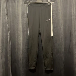 Svarta Nike Dri-FIT träningsbyxor M - Svarta träningsbyxor från Nike med Dri-FIT-teknologi. Byxorna har vita sidoränder, elastisk midja och smal passform. Perfekta för träning eller chill, med klassisk Nike-logga på låret. Materialet är syntet för bästa komfort och andningsförmåga.