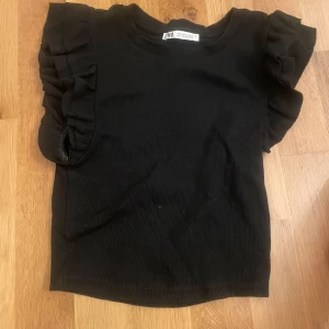 Svart topp med volangärm - Svart ribbad topp från Zara med korta ärmar och snygga volangdetaljer på axlarna. I storlek S men passar mer som en XS. Enkel rund halsringning och mjukt material. Supersöt och passar till allt!!!🤩🤩(Tröjan är inte nopprig eller smutsig bliden blev inte så bra😔)