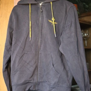 Svart hoodie Swedish Air Force South West - Svart hoodie med dragkedja från South West, Swedish Air Force-tryck i gult på ryggen och bröstet. Gula snören i huvan och känguruficka framtill. Perfekt för dig som gillar militärstil och vill sticka ut med coola detaljer. Köptes för 800