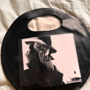 Svart rund handväska med foto-tryck - Unik rund handväska i svart skinnimitation med inbyggt handtag. Framsidan pryds av ett coolt svartvitt foto-tryck av en person med hatt och cigarett, vilket ger väskan en edgy och konstnärlig vibe. Perfekt accessoar för dig som vill sticka ut. Det är en liten klisterfläk som går att tvätta bort.