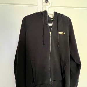 Svart hoodie från BOSS med dragkedja - Svart hoodie från BOSS i storlek M med guldfärgad BOSS-logga på bröstet. Tröjan har huva med snörning, hel dragkedja framtill och två fickor. Perfekt för chill dagar eller när du vill ha en clean och stilren look.