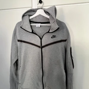 Grå hoodie från Nike med dragkedja - Säljer en grå hoodie från Nike i storlek L. Tröjan har hel dragkedja, huva och svarta detaljer längs bröstet och ärmen. Nike-loggan är broderad på vänster sida. Perfekt för chill eller träning, riktigt skön och stilren.