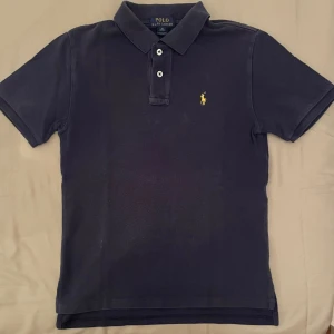  Ralph Lauren pike  - Klassisk pikétröja i mörkblått med Polo Ralph Lauren samt en gul logga. Perfekt både till vardag och festligare tillfällen.