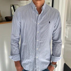 Randig skjorta från Ralph Lauren - Klassisk blå- och vitrandig skjorta från Ralph Lauren med button-down krage och broderad logga på bröstet. Skjortan har lång ärm, knappar framtill och är tillverkad i mjuk bomull. Perfekt för en clean och stilren look.