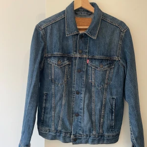 Levi's blå jeansjacka i storlek S - Klassisk blå jeansjacka från Levi's med snygga kontrastsömmar, två bröstfickor med knappar och raka linjer. Jackan har en tidlös design med krage och knäppning framtill. Perfekt för dig som vill ha en ikonisk denimlook.