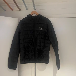 Svart dunjacka från EA7 Emporio Armani - Snygg svart dunjacka från EA7 Emporio Armani med quiltad design och huva. Jackan har dragkedja framtill, två fickor. Perfekt för kyliga dagar och enkel att matcha med allt. Klassisk och stilren look. De ända är en svart tyg fixning därför priset är som de e