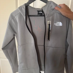 Grå vindjacka från The North Face - Grå dam-hoodie från The North Face med huva och dragkedja framtill. Storlek xs