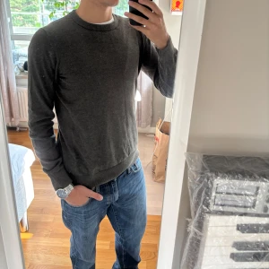 Mörkgrå stickad tröja herr - Snygg mörkgrå stickad tröja med rund hals och långa ärmar. Enkel och stilren design som passar till jeans eller chinos. Mjuk och skön känsla, perfekt för kyliga dagar. Tröjan har en normal passform och ribbade muddar vid ärmslut och nederkant.