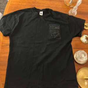 Svart Nike t-shirt cactusjack - Svart t-shirt från Nike med cool ficka på bröstet och liten orange swoosh. Klassisk passform och rund halsringning. På ryggen finns en diskret broderad text i orange. Perfekt för dig som gillar streetwear och snygga detaljer.